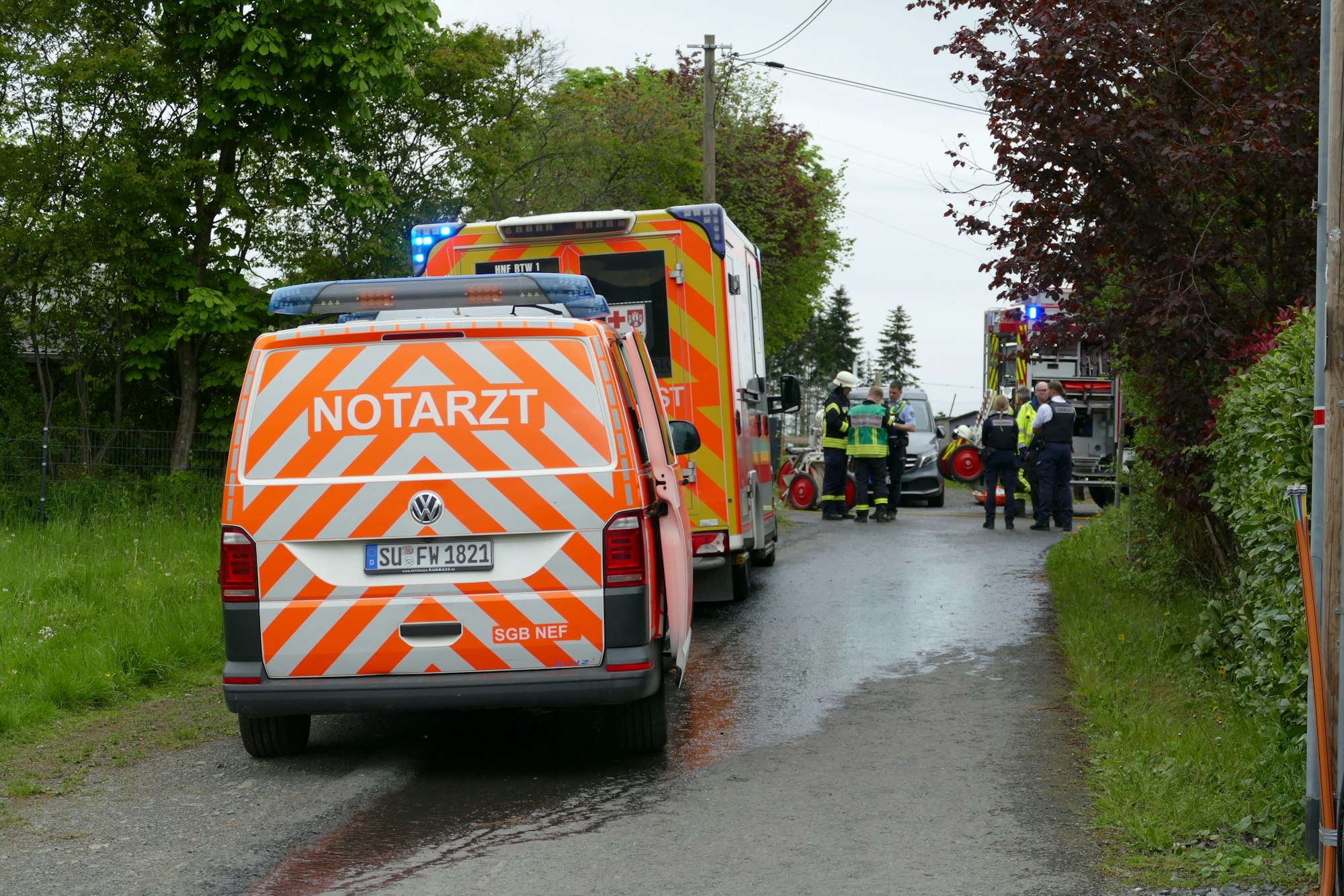 Ein Notarztwagen und ein Krankenwagen stehen auf einer asphaltierten Zufahrt. Im Hintergrund sind Feuerwehrleute und Rettungskräfte.