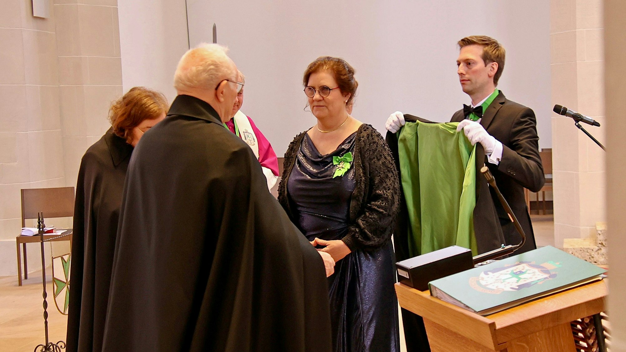 Inge Hartmann steht vor ihrem Vorgesetzten und kriegt einen grün-schwarzen Mantel umgelegt.