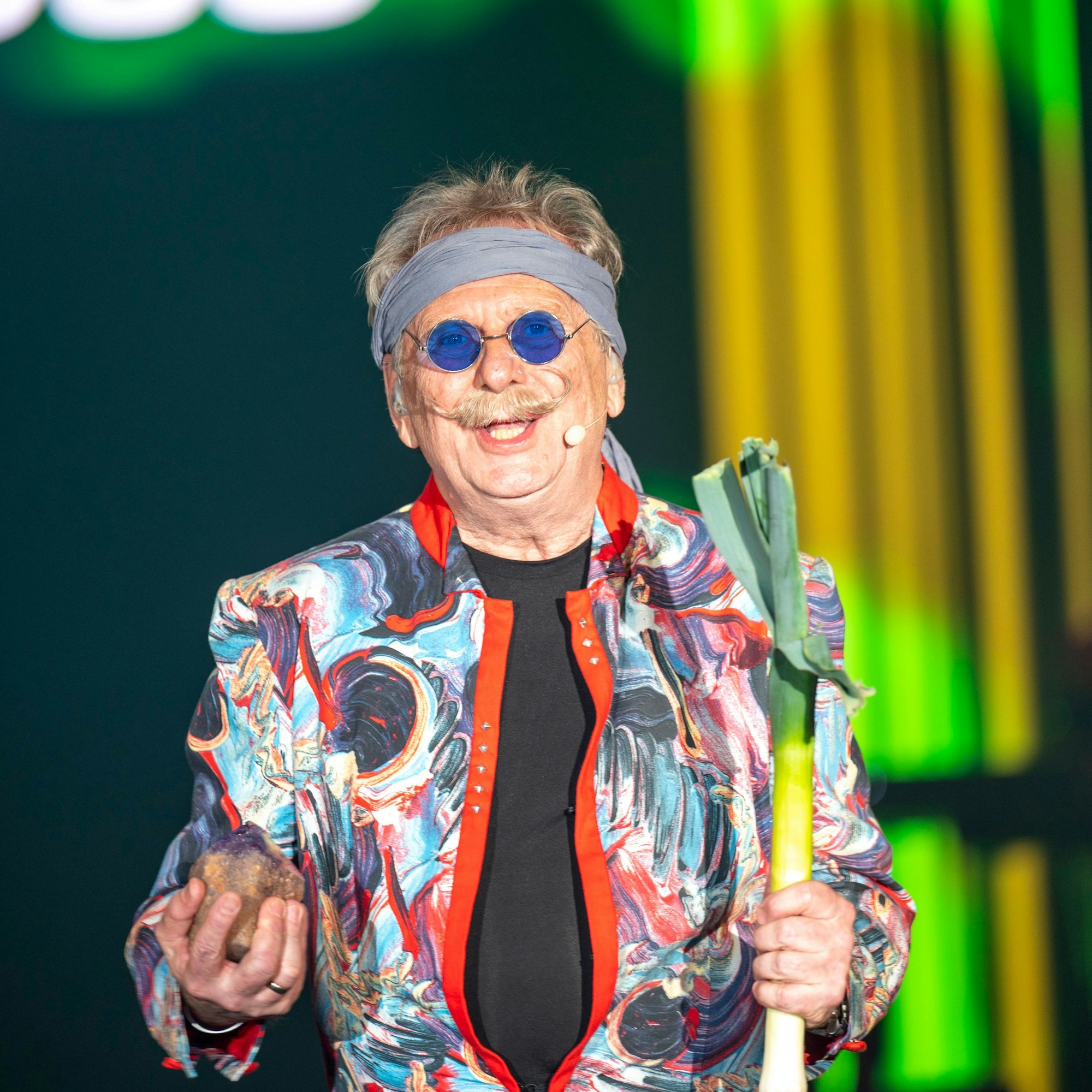 Henning Krautmacher hält einen Porree und einen Kristall in der Hand, dazu hat er eine blaue Brille auf und trägt ein buntes Jacket.