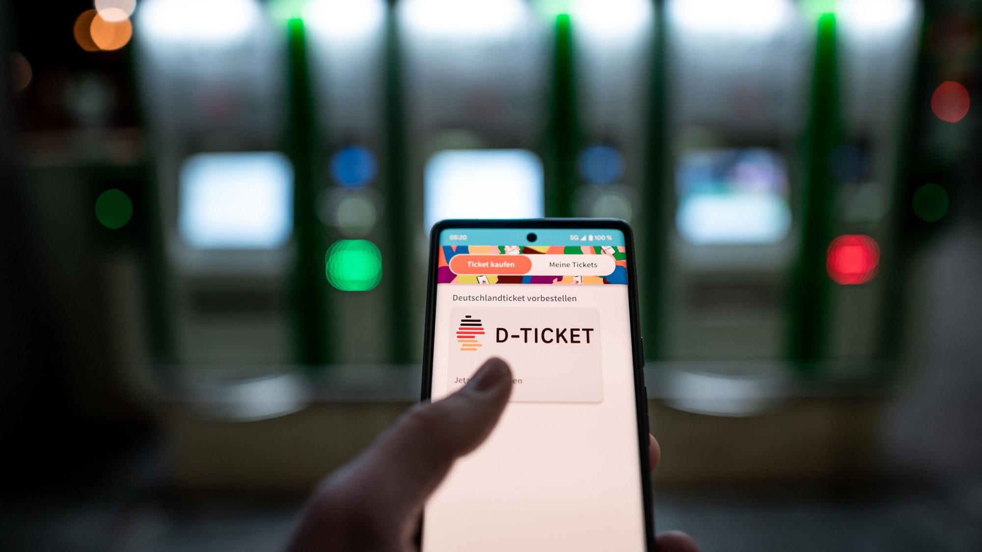 ARCHIV - 03.04.2023, Nordrhein-Westfalen, Essen: Ein Mann hält ein Mobiltelefon, auf dem das «D-Ticket» gekauft werden kann, vor Ticketautomaten am Hauptbahnhof Essen in der Hand (zu dpa: «Schüler, Eltern und GEW kritisieren 49-Euro-Ticket als ungerecht»). Foto: Fabian Strauch/dpa +++ dpa-Bildfunk +++
