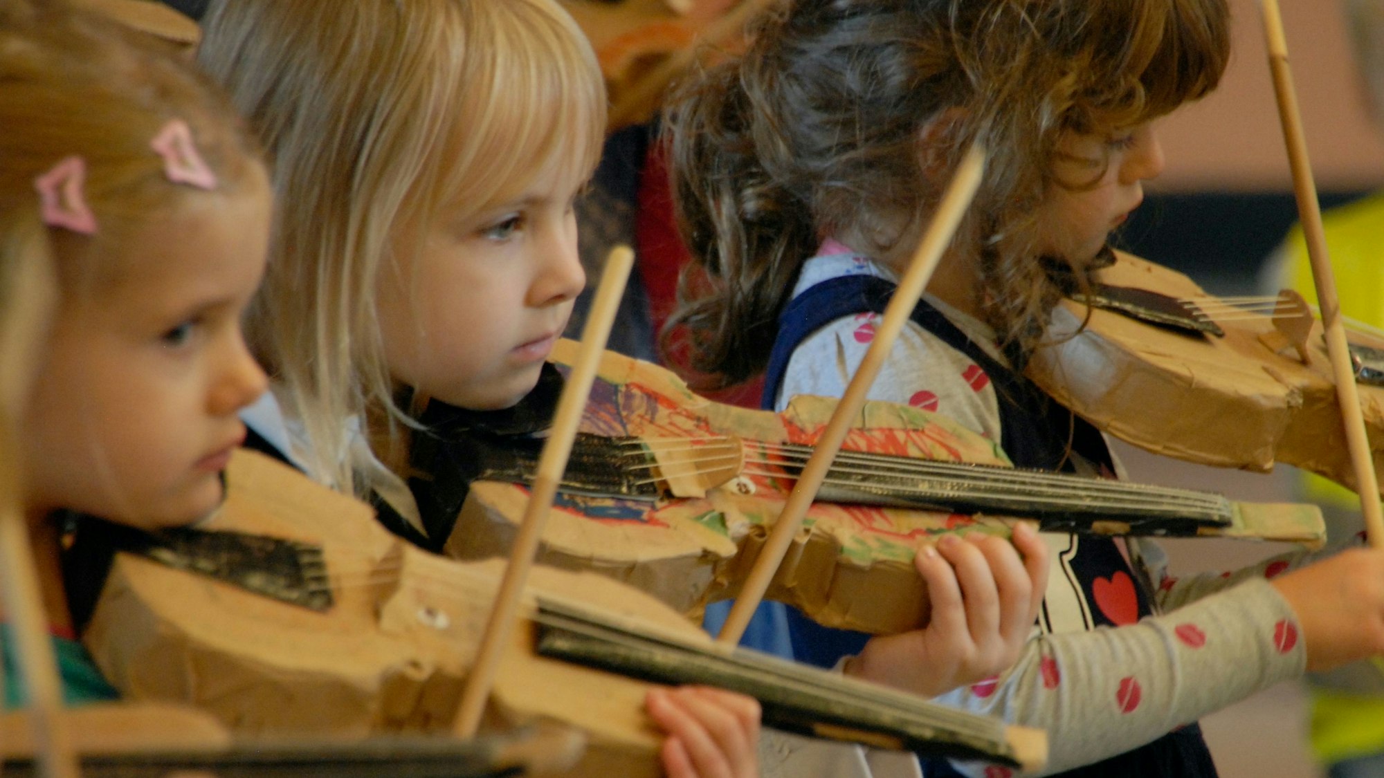Drei Mädchen im Grundschulalter spielen auf einer Geige aus Pappe