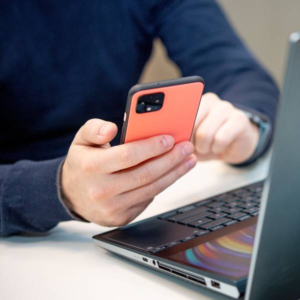Eine Person sitzt am Schreibtisch vor einem Laptop und hält dazu das Smartphone in der Hand.