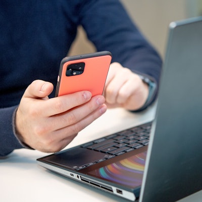 Eine Person sitzt am Schreibtisch vor einem Laptop und hält dazu das Smartphone in der Hand.