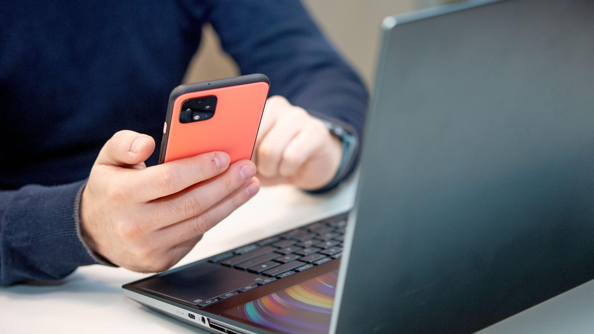 Eine Person sitzt am Schreibtisch vor einem Laptop und hält dazu das Smartphone in der Hand.