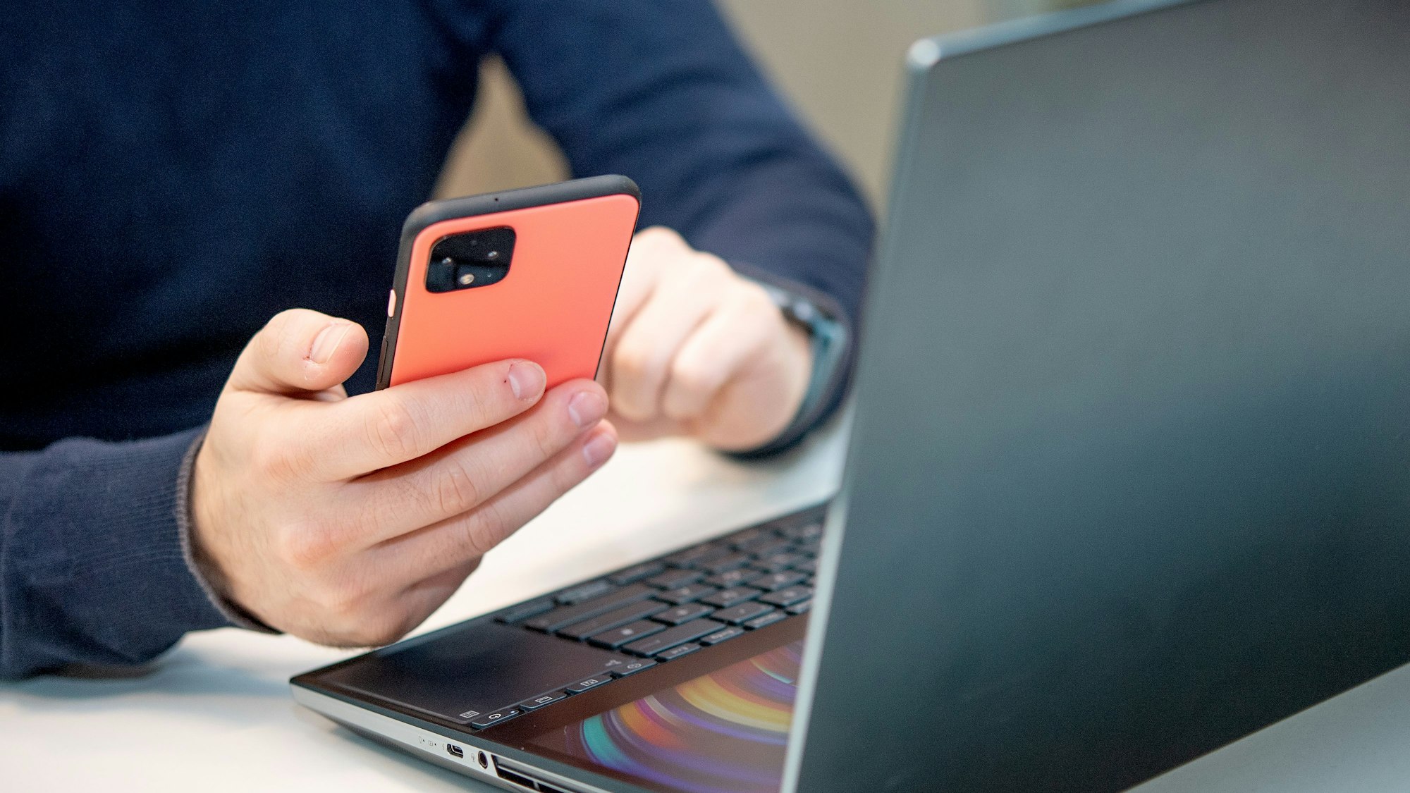 Eine Person sitzt am Schreibtisch vor einem Laptop und hält dazu das Smartphone in der Hand.