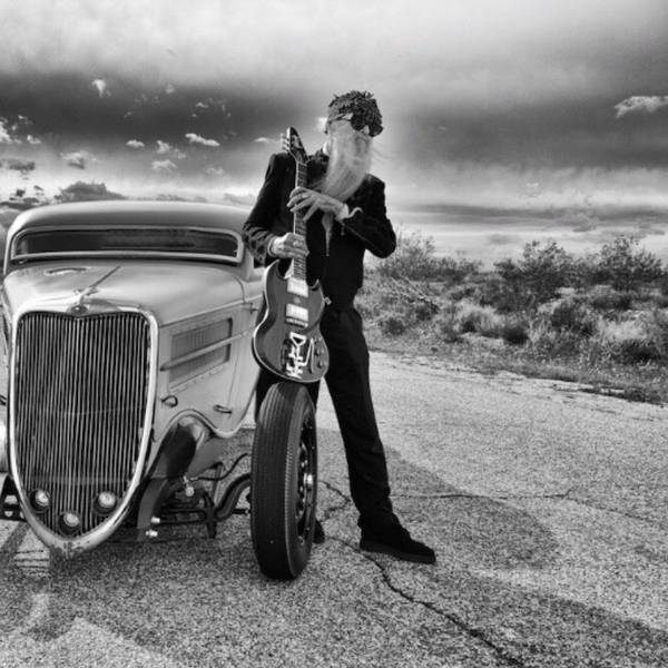 ZZ-Top-Gitarrist Billy F. Gibbons steht neben seinem Hot-Rod-Auto auf einer Wüstenstraße in den USA. In den Händen hält er seine E-Gitarre, aufgestützt auf einem Vordereifen des Wagens.
