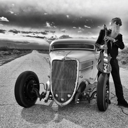 ZZ-Top-Gitarrist Billy F. Gibbons steht neben seinem Hot-Rod-Auto auf einer Wüstenstraße in den USA. In den Händen hält er seine E-Gitarre, aufgestützt auf einem Vordereifen des Wagens.