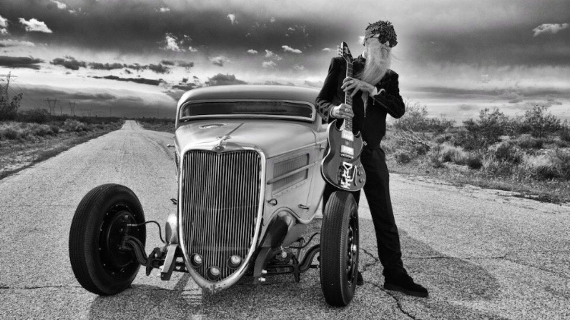 ZZ-Top-Gitarrist Billy F. Gibbons steht neben seinem Hot-Rod-Auto auf einer Wüstenstraße in den USA. In den Händen hält er seine E-Gitarre, aufgestützt auf einem Vordereifen des Wagens.