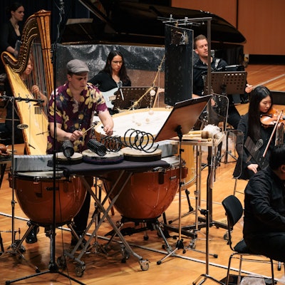 Koch sitzt am Schlagwerk, er trägt ein buntes Hemd. Weitere Musiker spielen auf der Bühne ihre Instrumente.