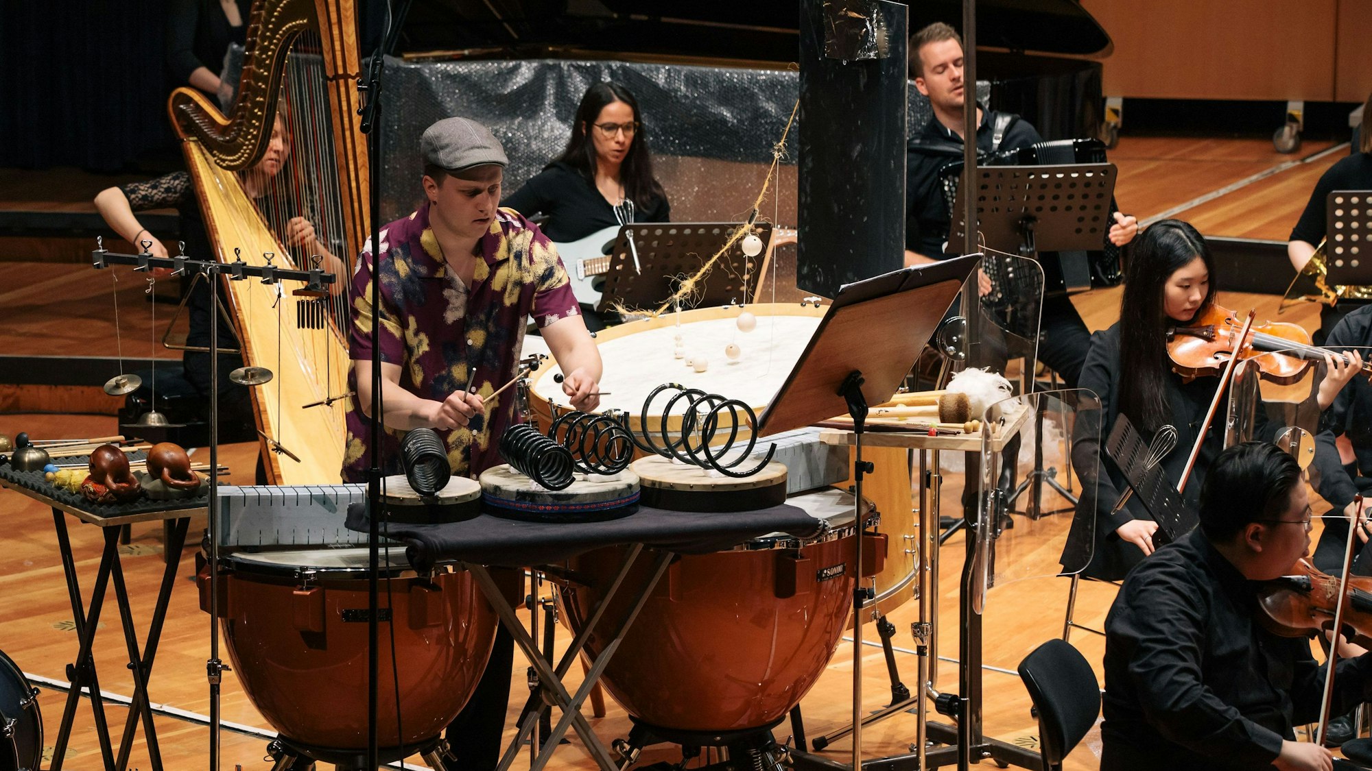Koch sitzt am Schlagwerk, er trägt ein buntes Hemd. Weitere Musiker spielen auf der Bühne ihre Instrumente.