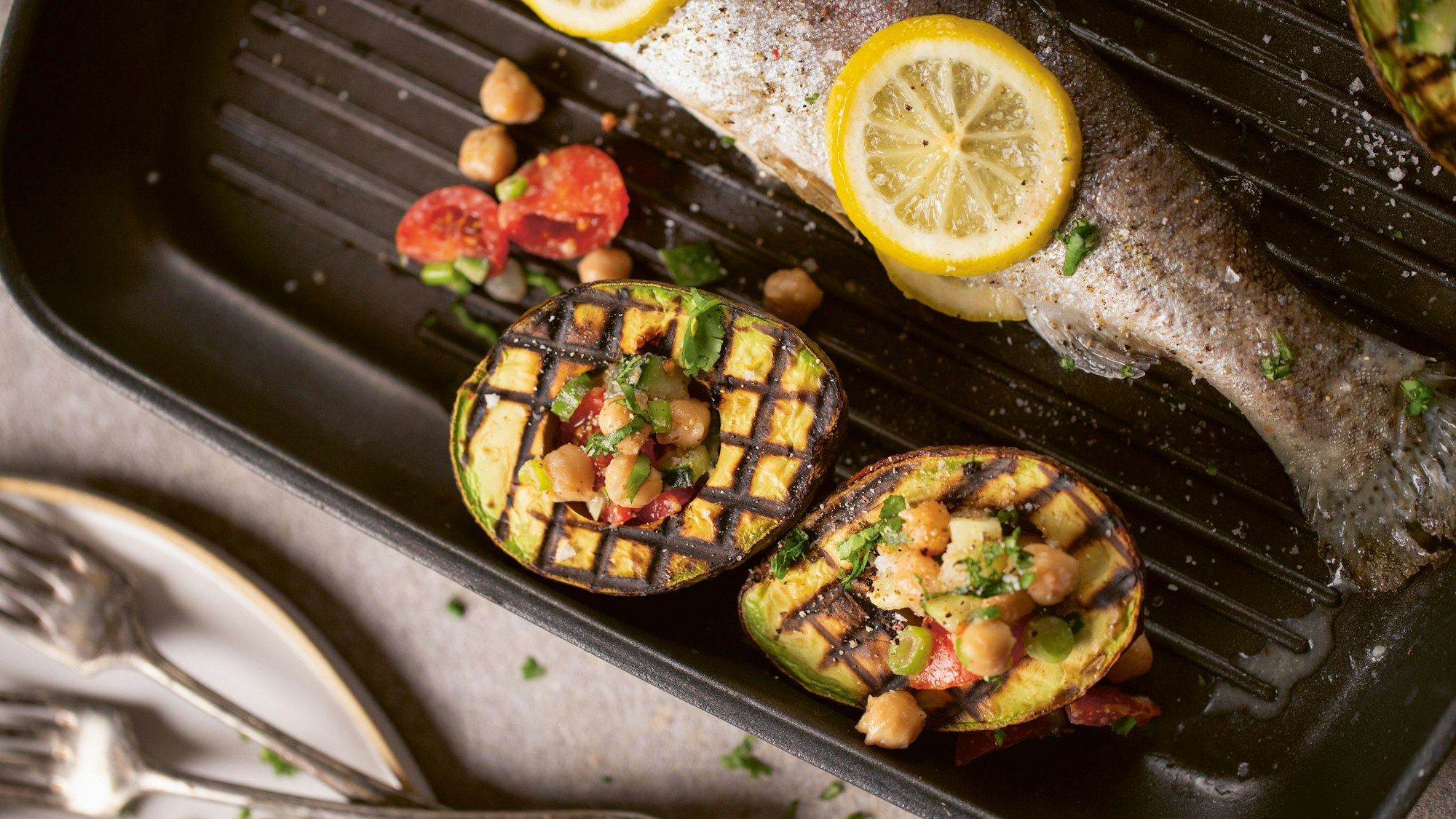 zwei gegrillte Avocado-Häften mit Füllung liegen neben einem gegrillten Fisch in einer Pfanne