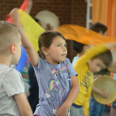 Ein kleines Mädchen schwingt eine Stofffahne, dahinter sieht man Kinder mit Trommeln.