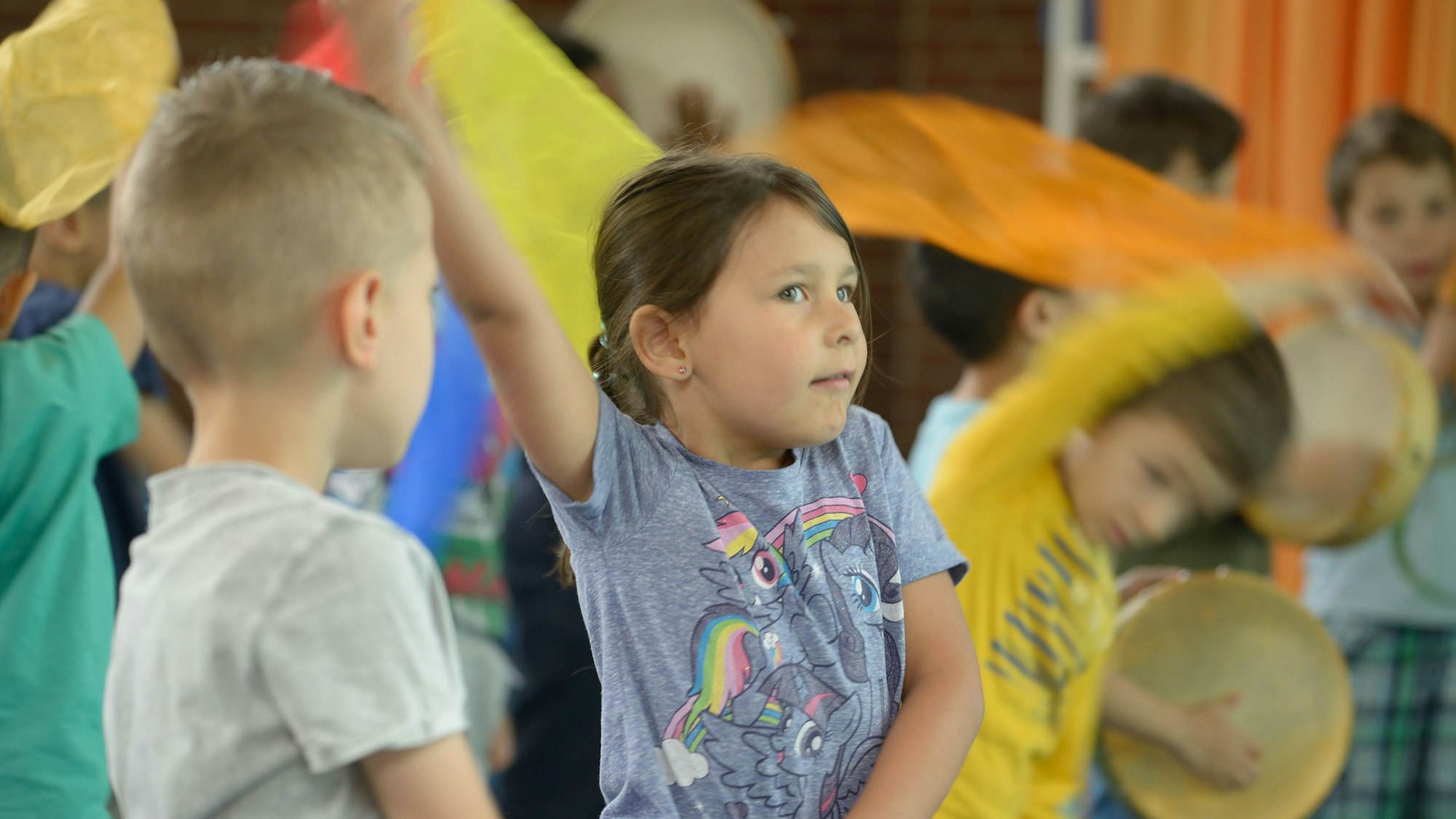 Ein Grundschulmädchen hält eine gelbe Stofffahne in die Luft und tanzt, es ist umgeben von Mitschülern, die auch Fahnen schwingen und sich begeistert bewegen.