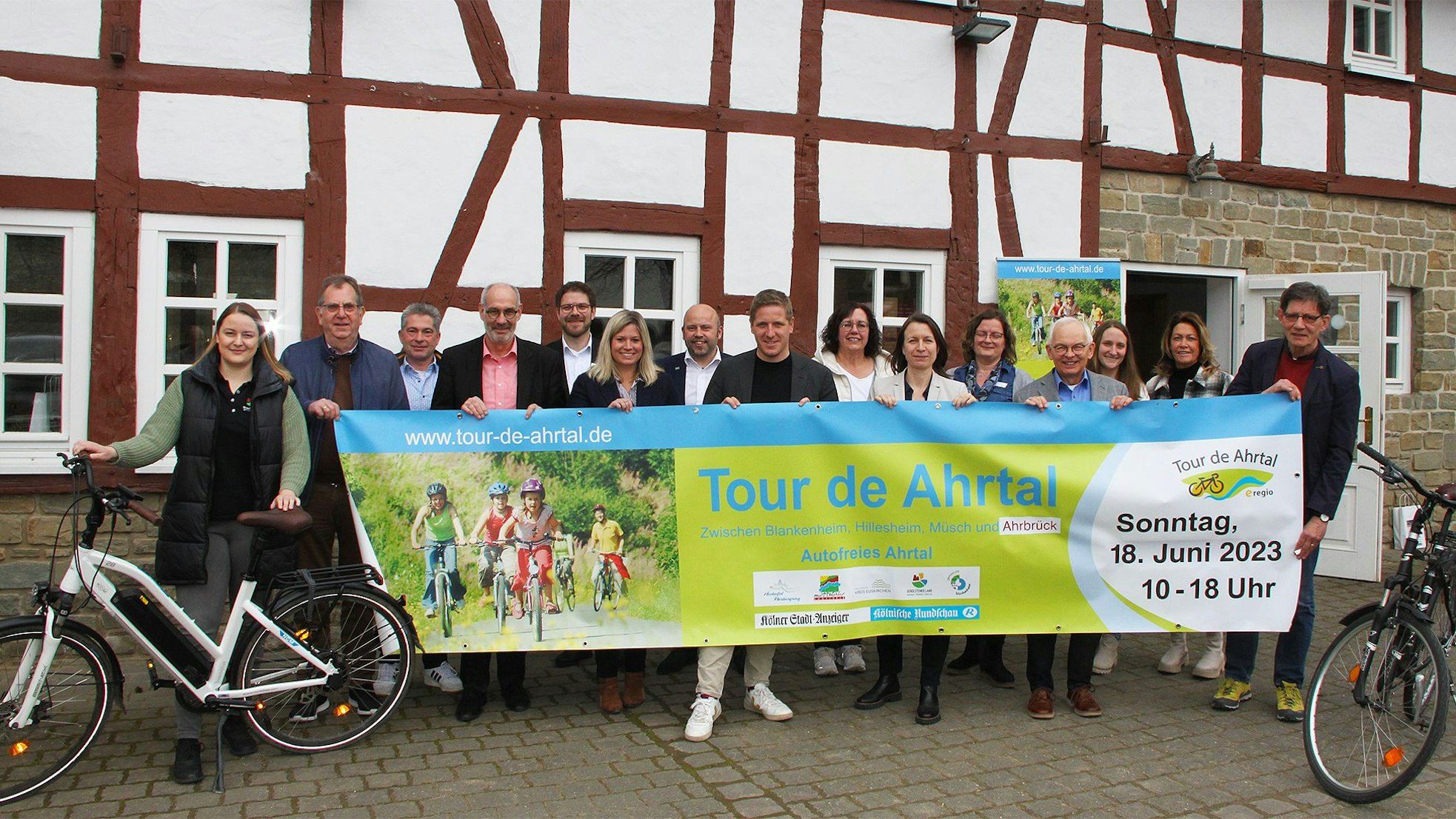 Die Vertreter der beteiligten Kreise und Kommunen halten vor einem Fachwerkgebäude in Antweiler ein Banner, mit dem für die „Tour de Ahrtal“ geworben wird.