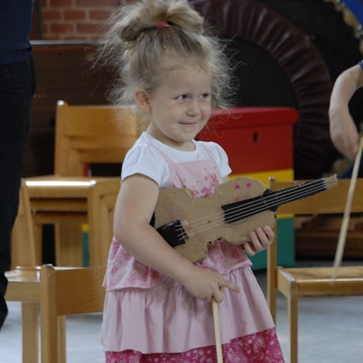 Ein kleines Mädchen mit rosafarbenem Kleid hält eine Geige aus Pappe unterm Arm.