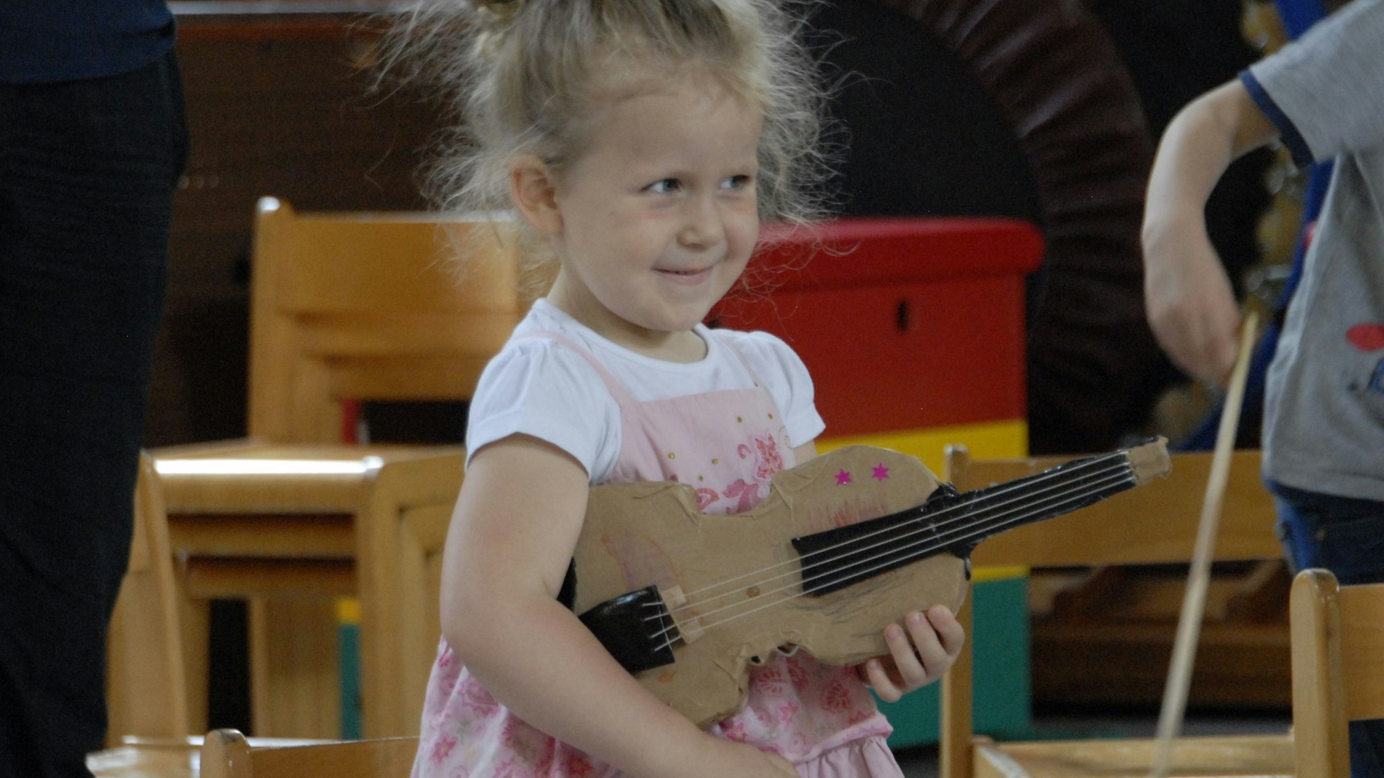 Ein kleines Mädchen mit rosafarbenem Kleid hält eine Geige aus Pappe unterm Arm.