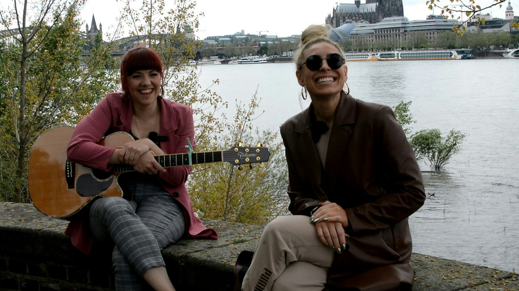 Johanna Eicken und Linda Teodosiu sitzen auf einer Mauer am Rhein, hinten ist der Dom zu sehen. Eicker hat eine Gitarre dabei.