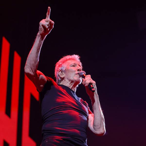 Roger Waters in der Lanxess Arena, er hält sich ein Mikro an den Mund, während er mit der rechten Hand aufzeigt.