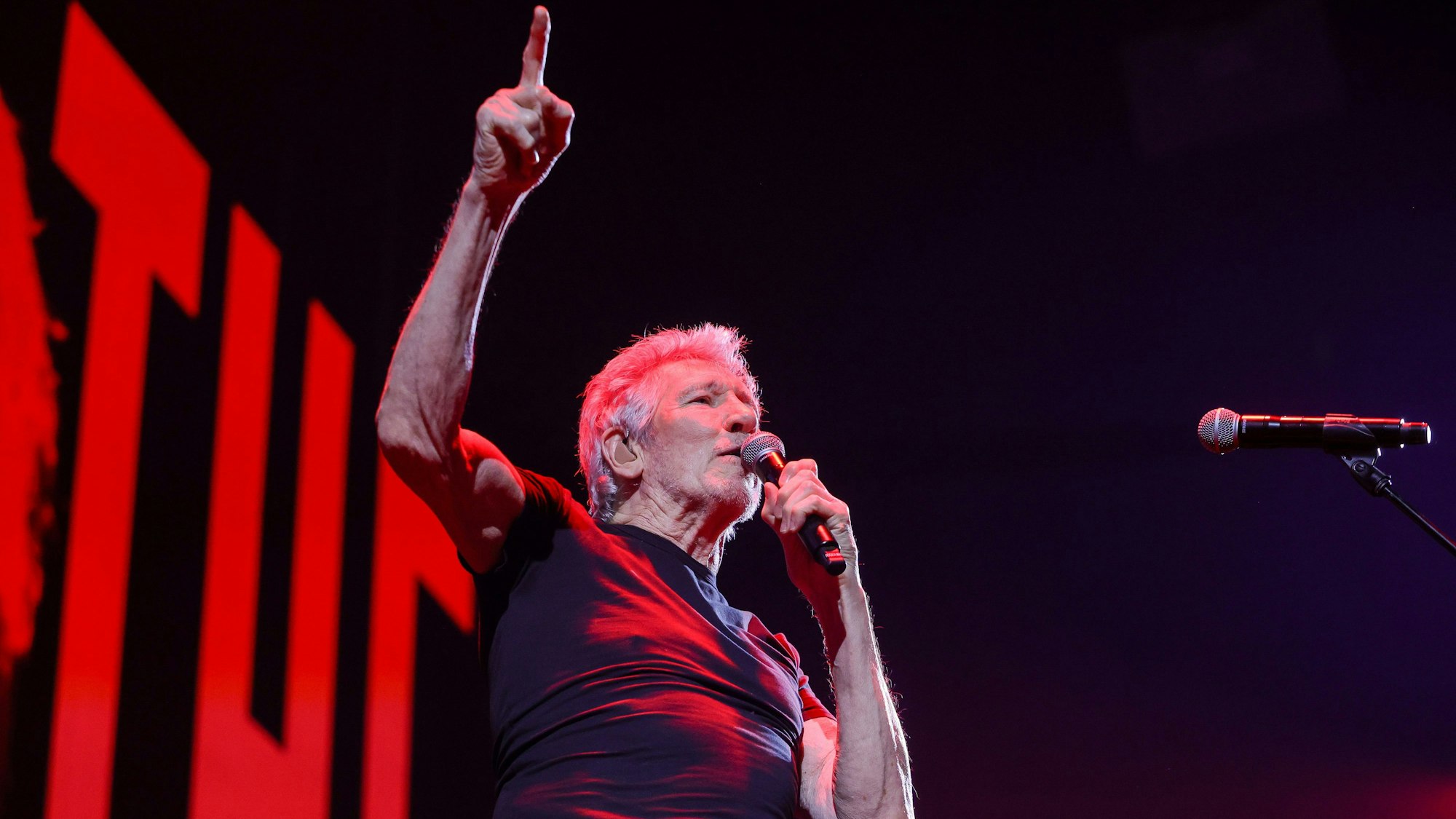 Roger Waters in der Lanxess Arena, er hält sich ein Mikro an den Mund, während er mit der rechten Hand aufzeigt.