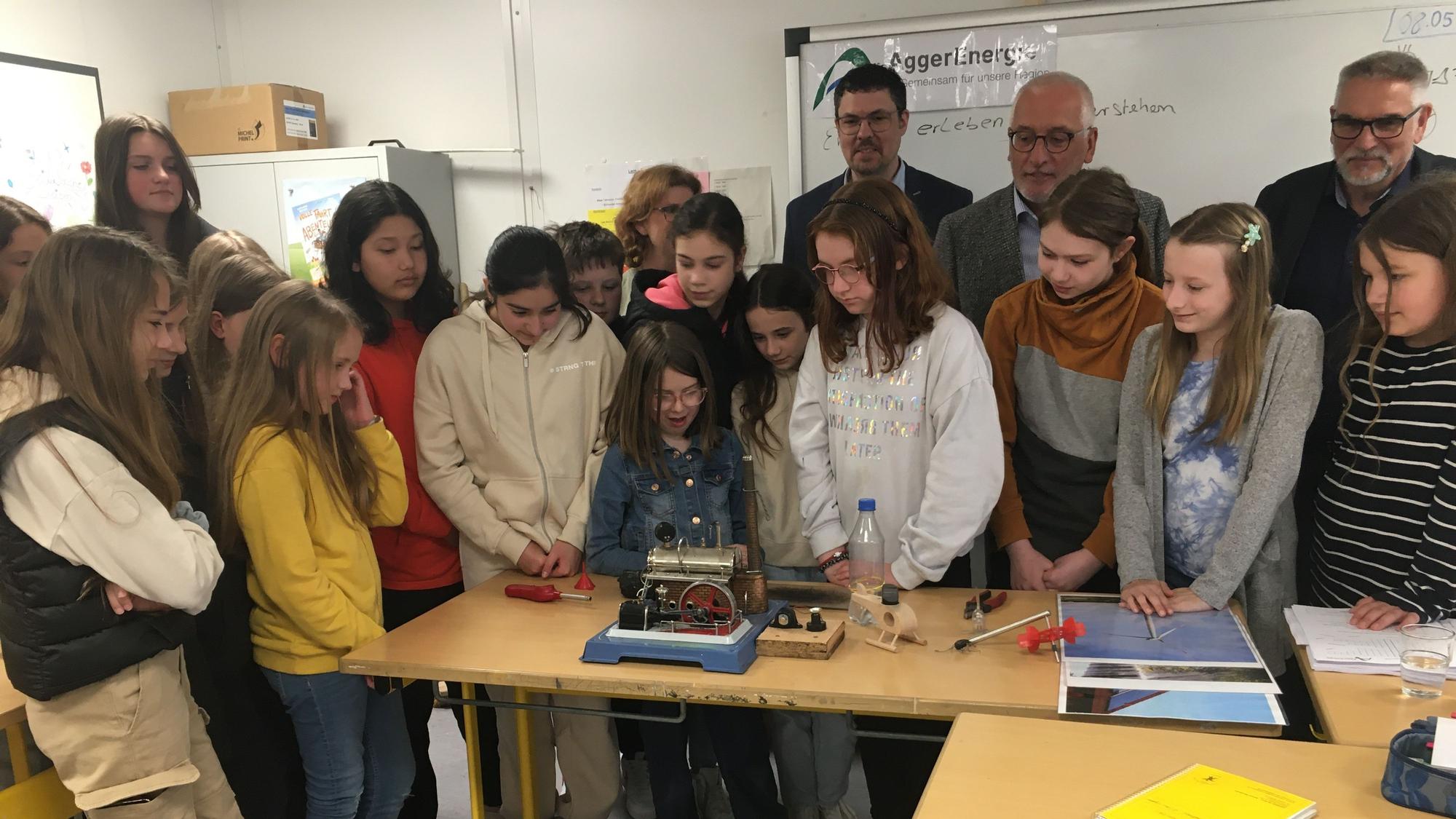 Eine Schulklasse steht als Gruppe neugierug vor einem Experiment, das auf dem Tisch vor ihr aufgebaut ist.