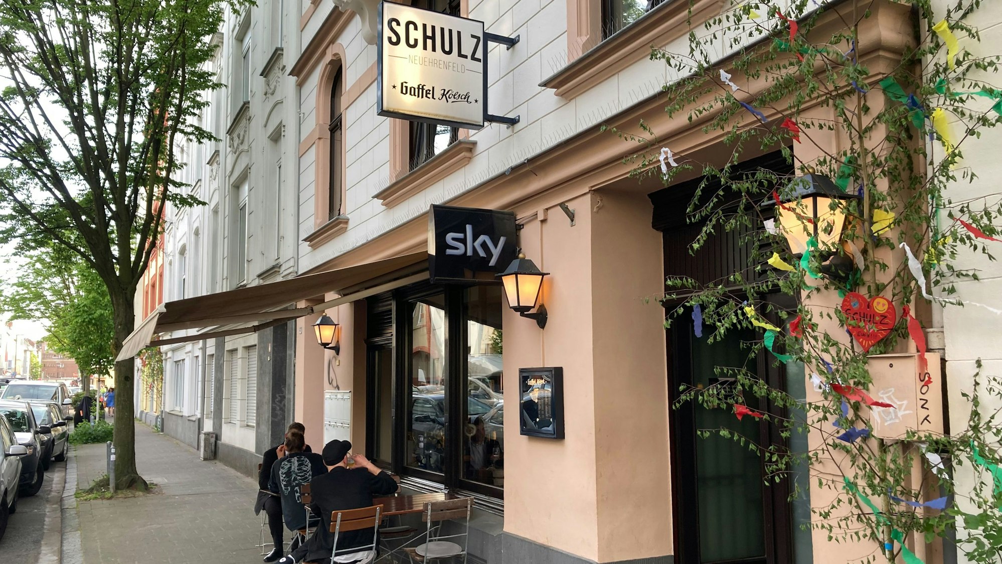 Außenansicht des Lokals „Schulz“ in der Landmannstraße in Neuehrenfeld: Ein geschmückter Maibaum ziert die rechte Seite des Eingangs zur Kneipe, auf deren Namen eine Leuchtreklame hinweist. Unter einem ausgezogenen Sonnenschutz stehen zwei Tische mit Stühlen, die von einigen Gästen genutzt werden.