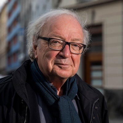 Der Journalist und Filmemacher Osman Okkan mit Brille, Hemd, Schal und Jacke und einer Kölner Straße im Hintergrund.