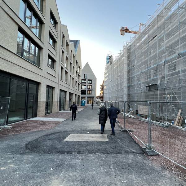 In der Porzer Innenstadt befindet sich links ein vierstöckiges, neues Gebäude, dessen Erdgeschoss für Ladenlokale vorgesehen ist. Am gegenüber liegenden Gebäude in gleicher Höhe wird noch gebaut, Schutzplanen verhängen die Sicht. Im Bildhintergrund ist ein ebenfalls neues Gebäude mit markantem Spitzdach zu sehen.