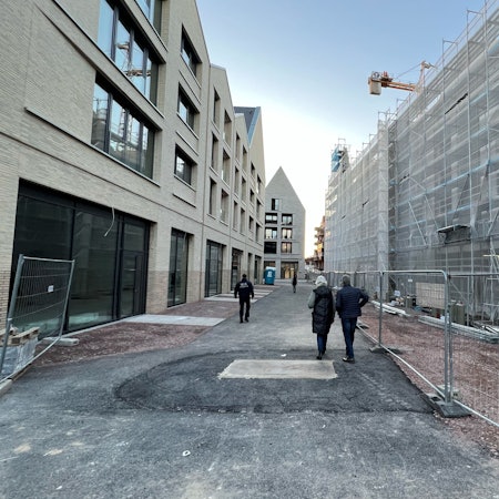 In der Porzer Innenstadt befindet sich links ein vierstöckiges, neues Gebäude, dessen Erdgeschoss für Ladenlokale vorgesehen ist. Am gegenüber liegenden Gebäude in gleicher Höhe wird noch gebaut, Schutzplanen verhängen die Sicht. Im Bildhintergrund ist ein ebenfalls neues Gebäude mit markantem Spitzdach zu sehen.