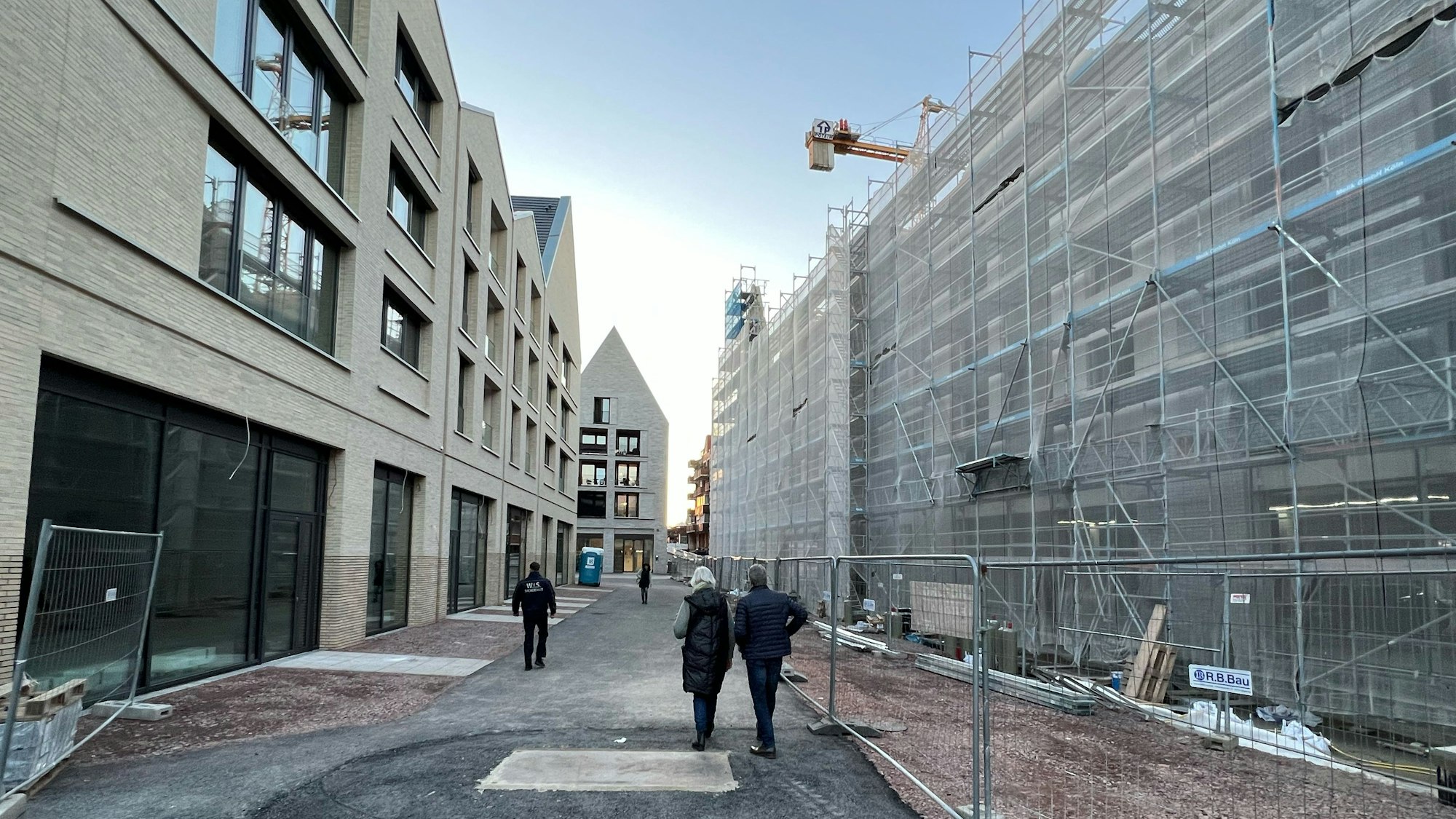 In der Porzer Innenstadt befindet sich links ein vierstöckiges, neues Gebäude, dessen Erdgeschoss für Ladenlokale vorgesehen ist. Am gegenüber liegenden Gebäude in gleicher Höhe wird noch gebaut, Schutzplanen verhängen die Sicht. Im Bildhintergrund ist ein ebenfalls neues Gebäude mit markantem Spitzdach zu sehen.