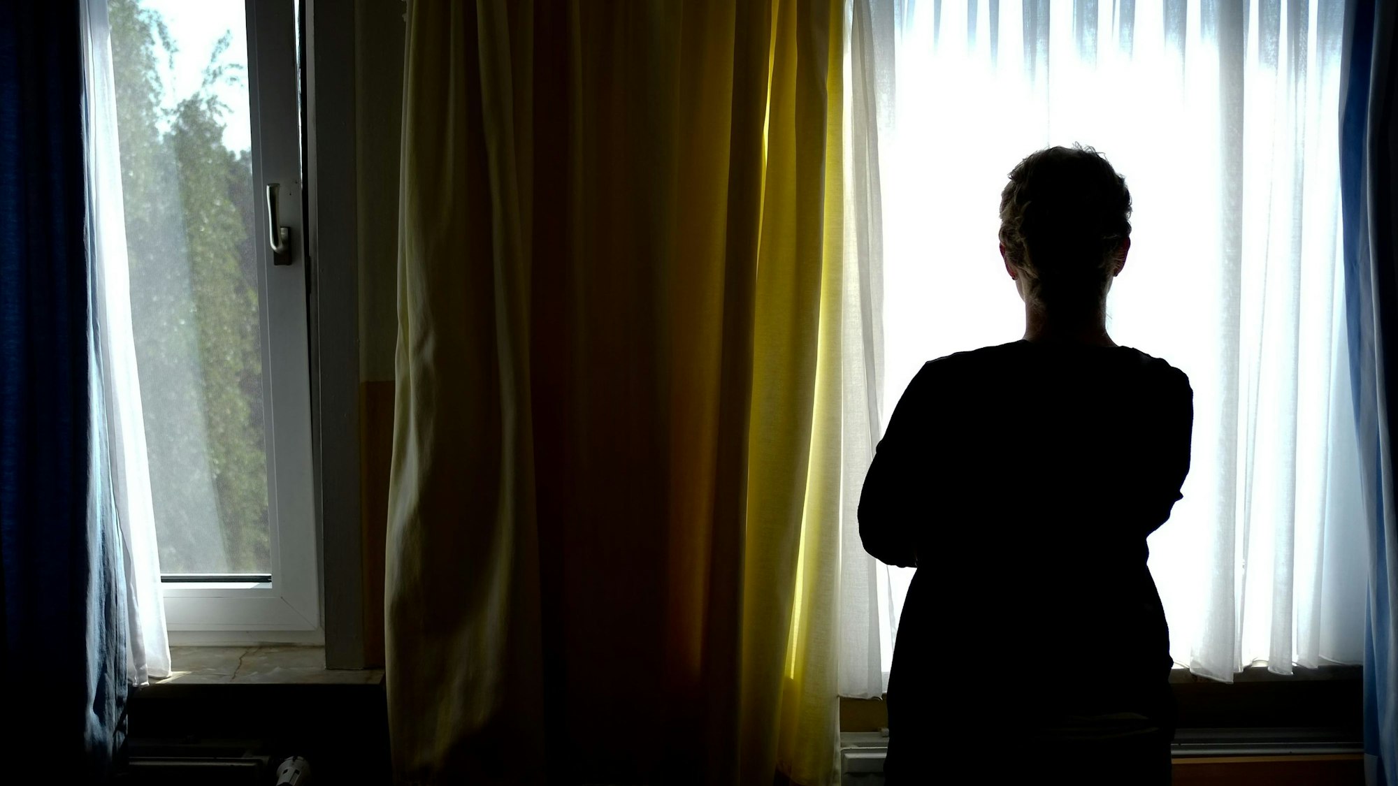 Eine Person steht vor einem Fenster und ist im Gegenlicht nur als dunkle Silhouette erkennbar.