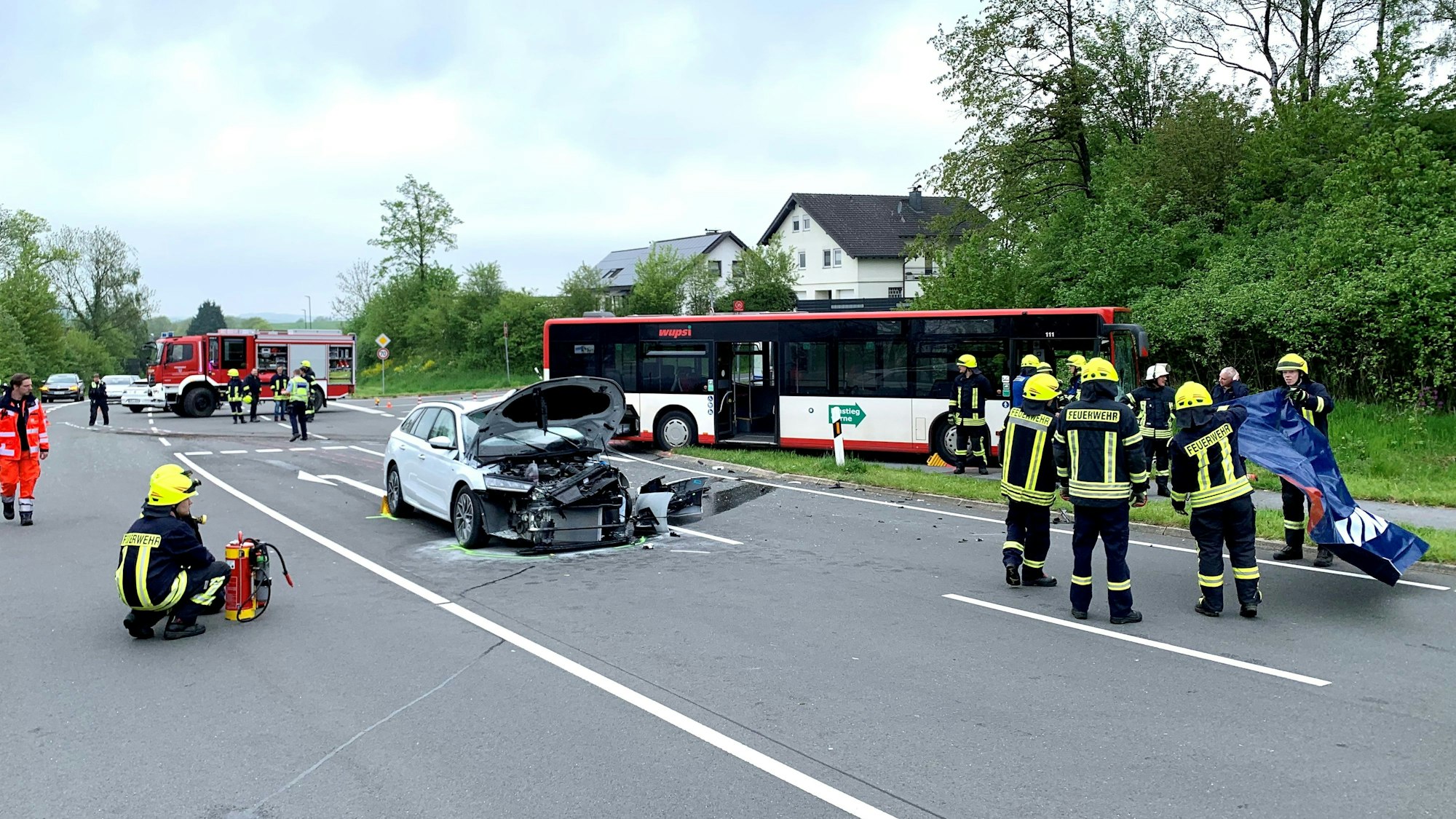 Der Pkw ist stark zerstört, die Motorhaube ist offen. Im Hintergrund der Bus. Einsatzkräfte der Feuerwehr stehen im Unfallbereich.