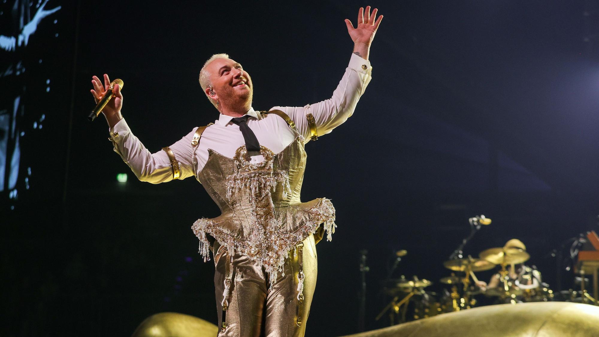 Sam Smith am 8. Mai 2023 in der Lanxess-Arena. Die nicht-binäre Person trägt ein goldenes Bustier über einem weißen Hemd und eine goldene Hose. Sie hält die Arme ausgestreckt und lächelt.