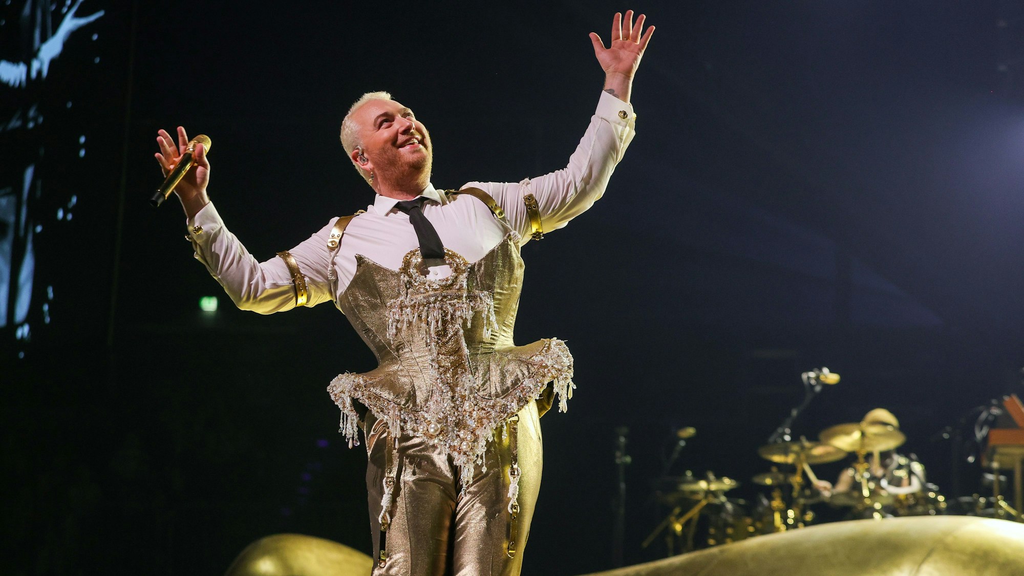 Sam Smith am 8. Mai 2023 in der Lanxess-Arena. Die nicht-binäre Person trägt ein goldenes Bustier über einem weißen Hemd und eine goldene Hose. Sie hält die Arme ausgestreckt und lächelt.