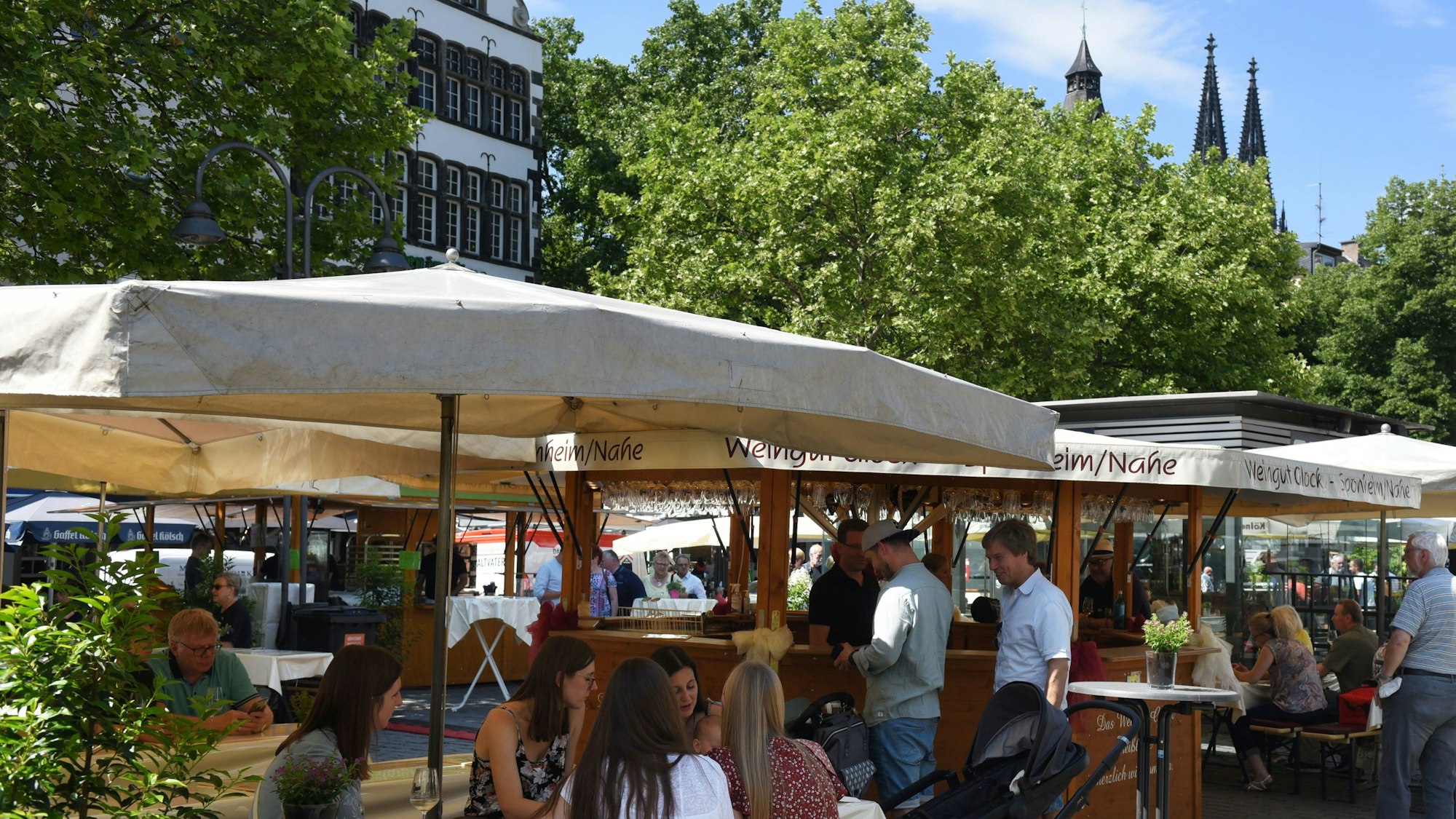 Sonnenschirme und Stände bei der Kölner Weinwoche auf dem Heumarkt. Davor stehen Menschen und trinken Wein.