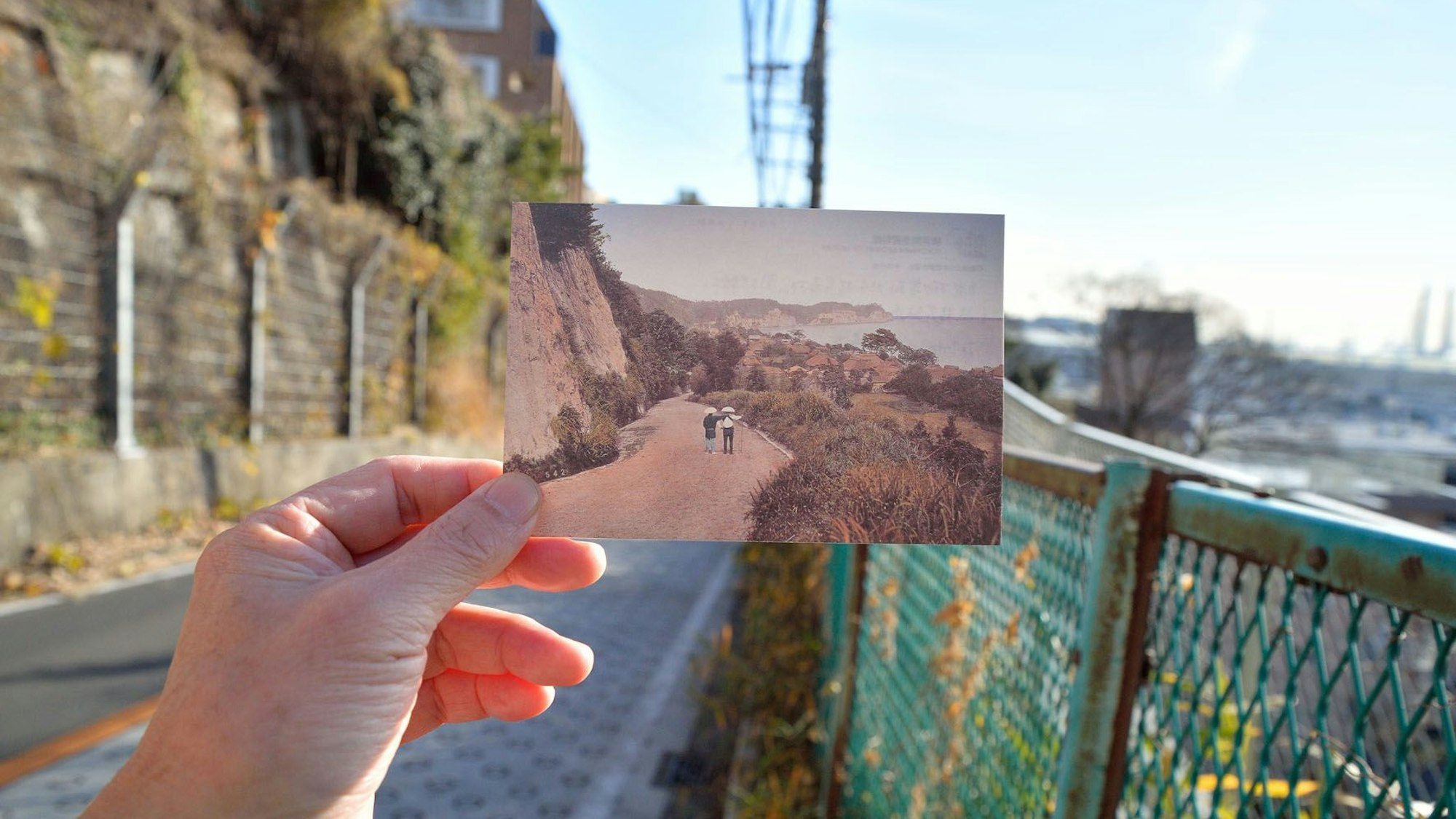 Jemand hält eine alte Postkarte ins Bild. Man erkennt, dass beide Aufnahmen einer Straße denselben Ort zeigen, nur im Abstand von über 100 Jahren.