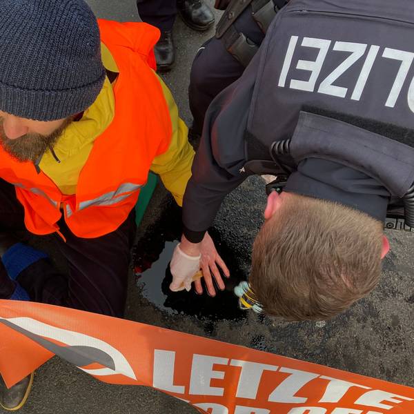Ein Klima-Aktivist sitzt auf einer Straße in Köln, nachdem die Letzte Generation in köln den morgendlichen Berufsverkehr stellenweise lahmlegten. Er hat sich an der Straße festgeklebt und hält ein Banner der Letzten Generation. Ein Polizist versucht, seine Hand von der Straße zu lösen.