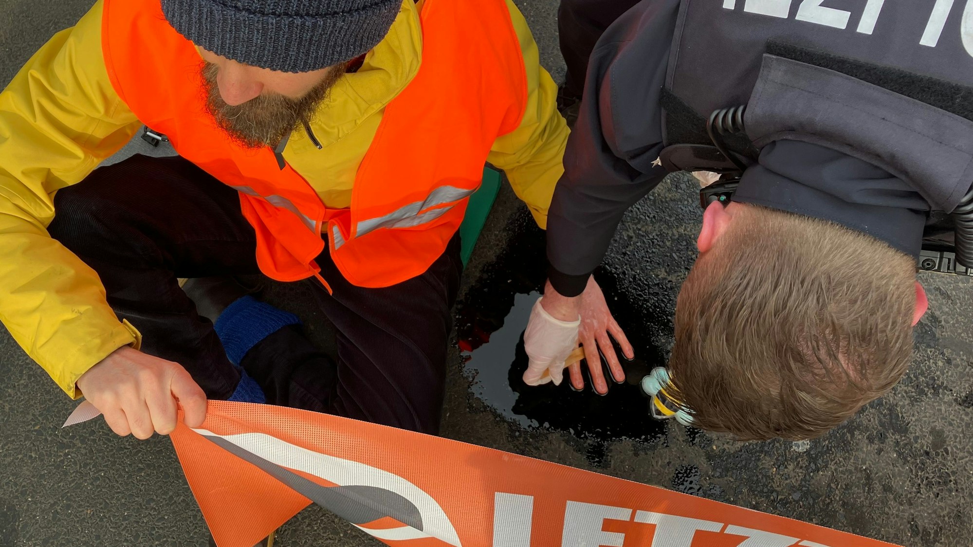 Ein Klima-Aktivist sitzt auf einer Straße in Köln, nachdem die Letzte Generation in köln den morgendlichen Berufsverkehr stellenweise lahmlegten. Er hat sich an der Straße festgeklebt und hält ein Banner der Letzten Generation. Ein Polizist versucht, seine Hand von der Straße zu lösen.