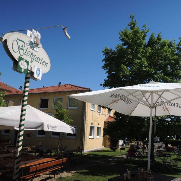 Vor einem Geböude stehen Tische, Stühle und Sonnenschirme. Auf einem Schild ist die Aufschrift „Biergarten“ zu lesen.