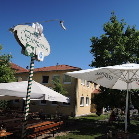 Vor einem Geböude stehen Tische, Stühle und Sonnenschirme. Auf einem Schild ist die Aufschrift „Biergarten“ zu lesen.