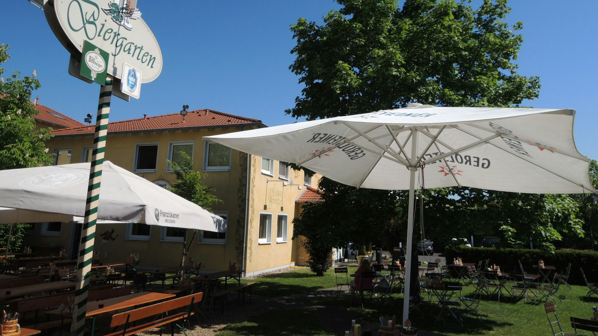 Vor einem Geböude stehen Tische, Stühle und Sonnenschirme. Auf einem Schild ist die Aufschrift „Biergarten“ zu lesen.