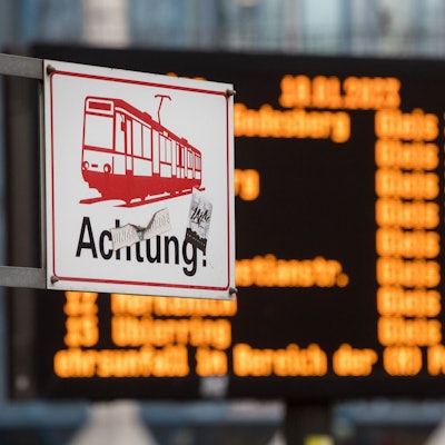 Das Bild zeigt ein Schild einer Straßenbahn mit dem Wort Achtung. Im Hintergrund sieht man eine verschwommene Anzeigetafel.