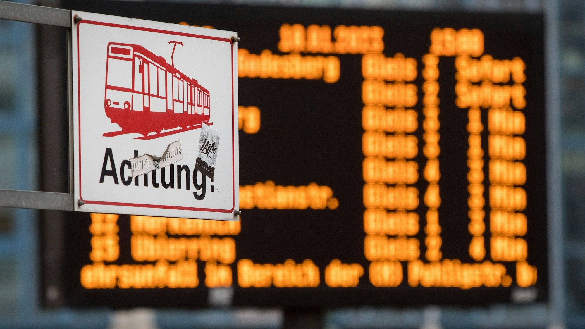 Das Bild zeigt ein Schild einer Straßenbahn mit dem Wort Achtung. Im Hintergrund sieht man eine verschwommene Anzeigetafel.