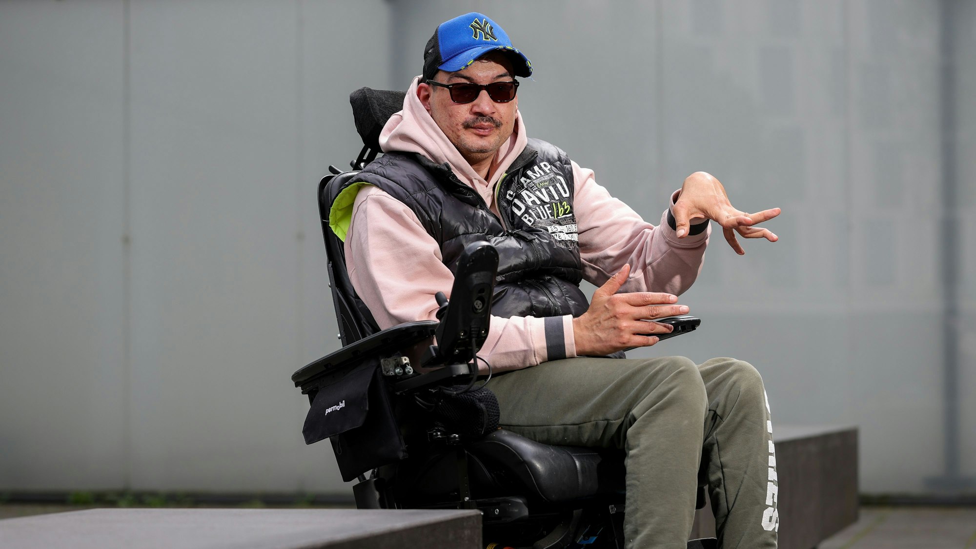 Rocky Klimt sitzt im Rollstuhl und schaut in die Kamera. Seine rechte Hand ist gehoben, sie ist durch seine spastische Lähmung verkrampft.