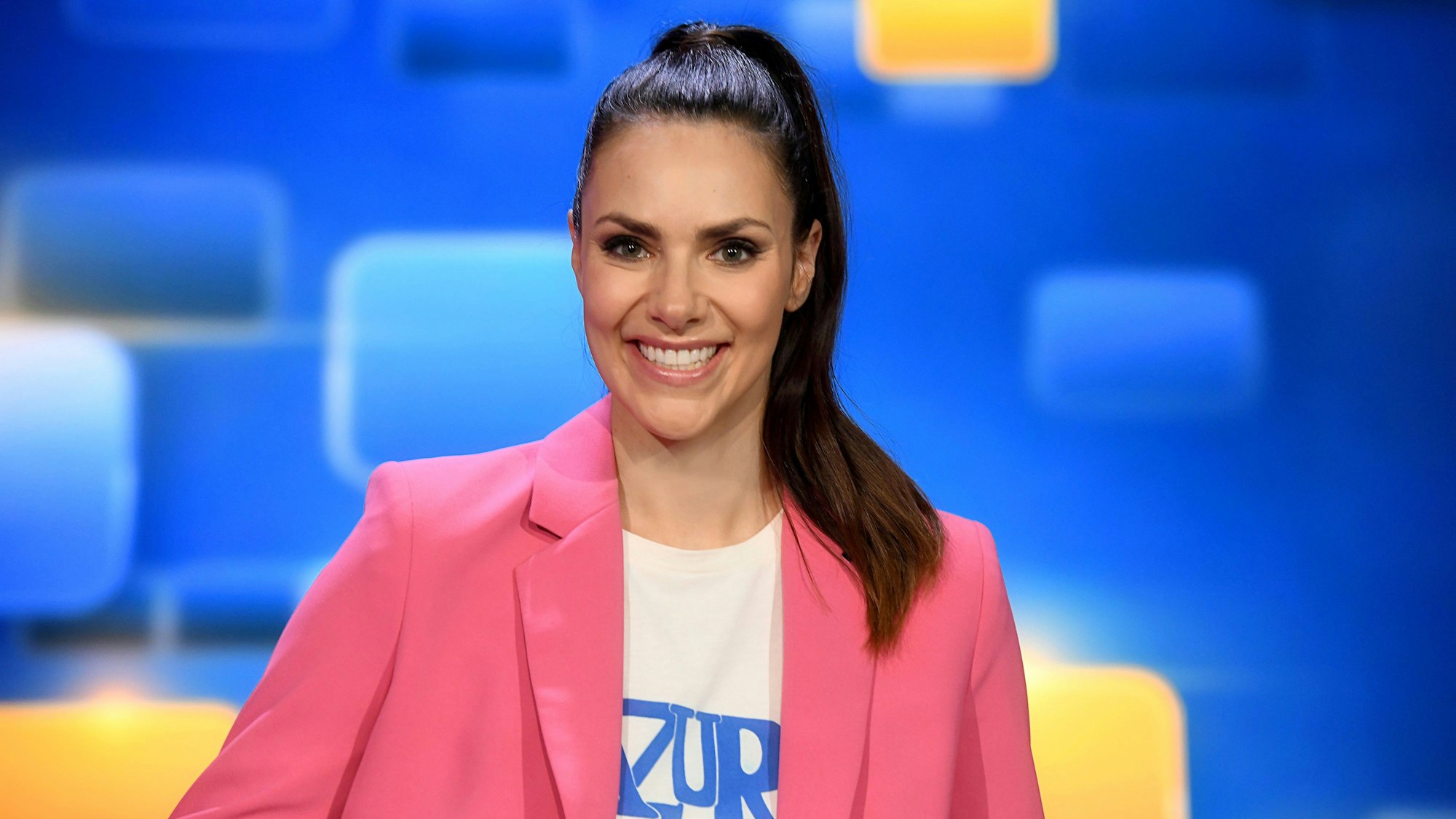 Moderatorin Esther Sedlaczek in pinkem Blazer mit weißem T-Shirt und hoch gebundenem Pferdeschwanz vor blauem Hintergrund.