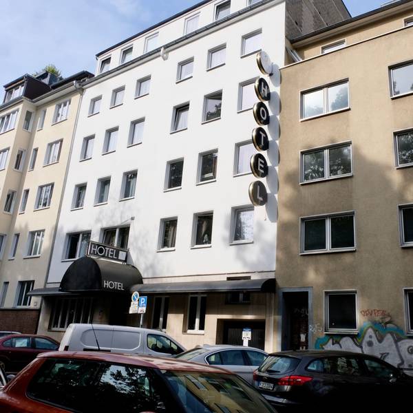 Eine Straße mit parkenden Autos ist zu sehen. Neben einem weiß getünchten Hotel ist ein vierstöckiges braunes Haus mit leeren Fenstern zu sehen.