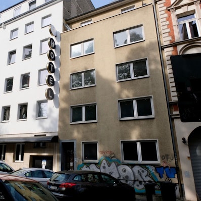 Eine Straße mit parkenden Autos ist zu sehen. Neben einem weiß getünchten Hotel ist ein vierstöckiges braunes Haus mit leeren Fenstern zu sehen.