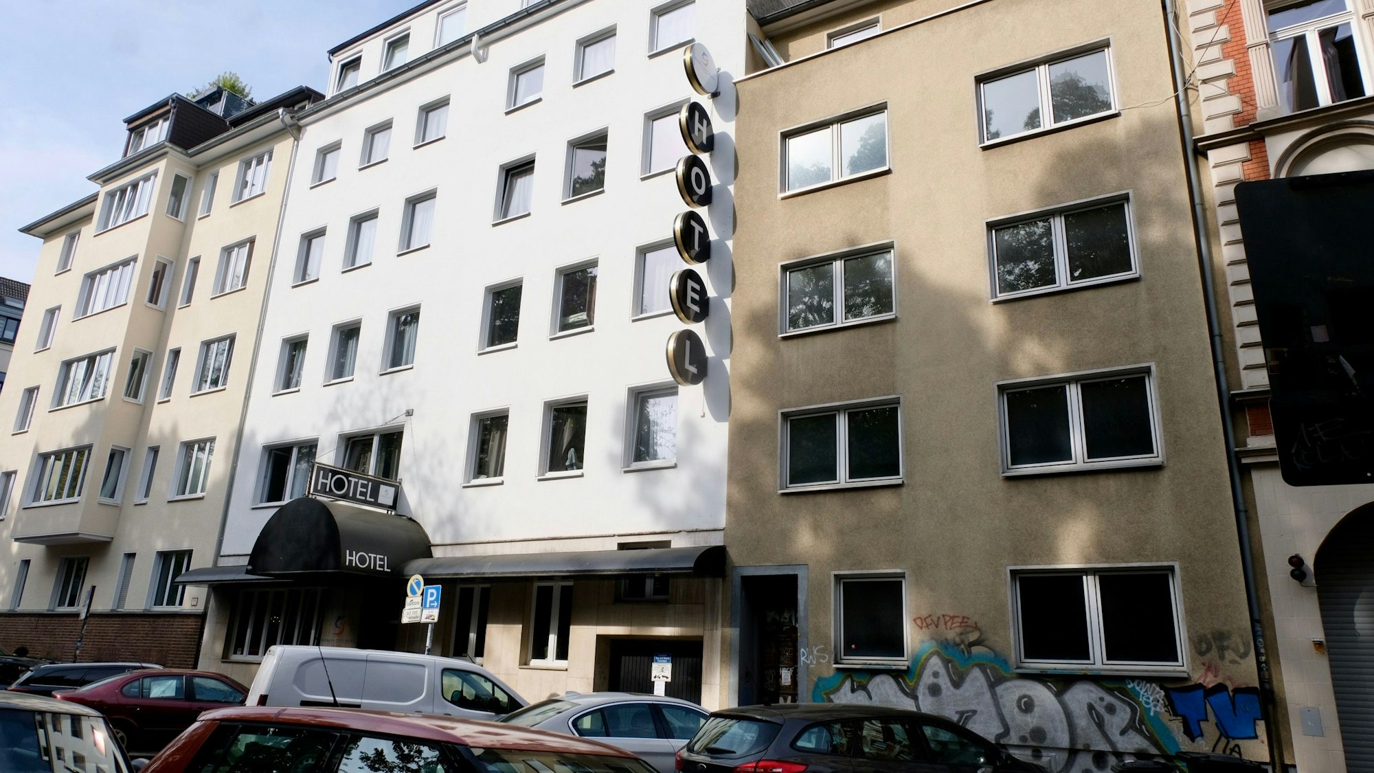 Eine Straße mit parkenden Autos ist zu sehen. Neben einem weiß getünchten Hotel ist ein vierstöckiges braunes Haus mit leeren Fenstern zu sehen.