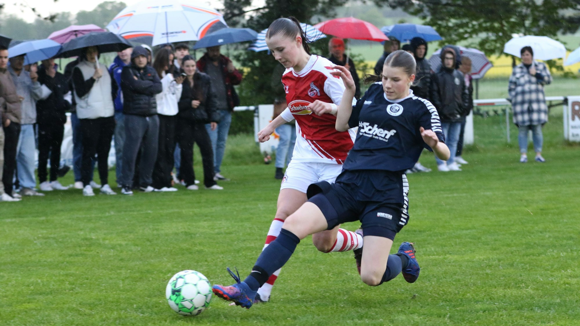 Eine Kommerner Spielerin setzt zu einer fairen Grätsche gegen eine Kölnerin an. Im Hintergrund stehen die Zuschauer mit Schirmen.