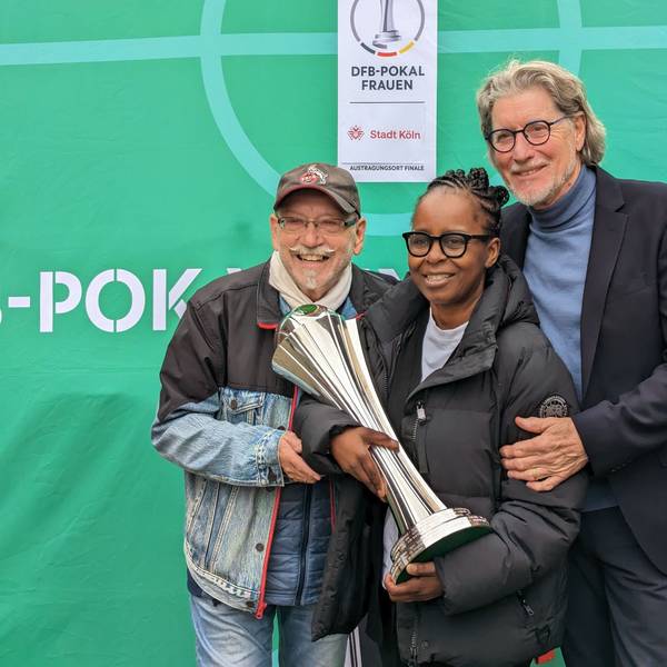 Janus Fröhlich, Shary Reeves und Toni Schumacher stehen zusammen vor einem grünen Hintergrund. Shary Reeves hat den Pokal in der Hand.