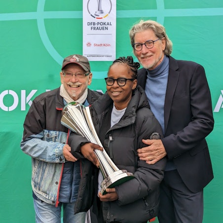 Janus Fröhlich, Shary Reeves und Toni Schumacher stehen zusammen vor einem grünen Hintergrund. Shary Reeves hat den Pokal in der Hand.